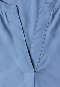 Gros plan d’un vêtement en tissu bleu à col en V avec des coutures visibles le long de l’encolure et de légers fronces près de la couture centrale.