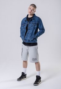 Jeansjack met klassieke snit, grijze shorts met zijprints en zwart-witte hoge sneakers met lichte accenten. Witte crew-sokken zichtbaar.