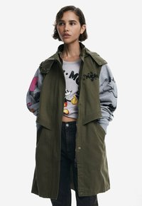 Manteau léger vert olive avec un col et plusieurs poches, porté sur un sweat-shirt gris avec des graphiques de dessins animés, complétant un jean noir.