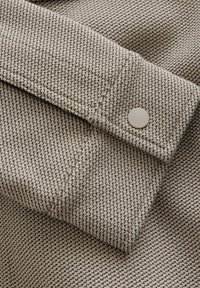 Poignet en tissu texturé beige et noir avec un seul bouton rond beige cousu près du bord.