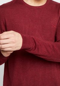 Punainen neulottu pusero pyöreällä pääntiellä, resorikädensuilla ja hillityllä logoyksityiskohdalla. Kangas vaikuttaa pehmeältä, kevyesti teksturoidulla pinnalla.