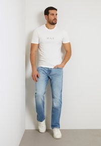 Valge lühikese varrukaga t-särk, millel on "GUESS" logo, koos helesiniste teksade ja valgete tossudega. Seisab neutraalse tausta ees.