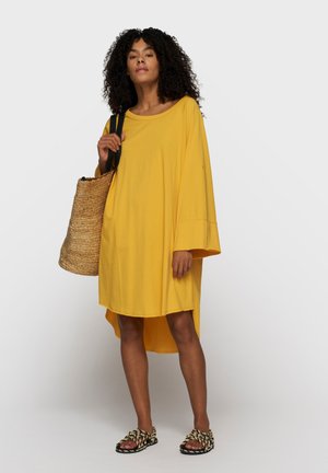 Kobieta w luźnej żółtej sukience, trzymająca plecioną torbę na ramię, ubrana w czarno-białe plecione sandały, stojąca na jednolitym tle.