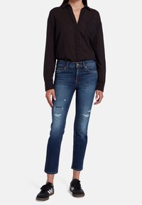 Camisa negra de botones, jeans azules desgastados con dobladillo deshilachado y zapatillas negras. La camisa tiene un bolsillo y los jeans tienen rotos.