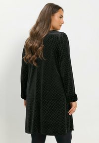 Schwarzes Samtkleid mit weißen Polka-Dots, langen Ärmeln und einer lockeren Passform. Es hat einen runden Ausschnitt und subtile Falten an den Schultern.