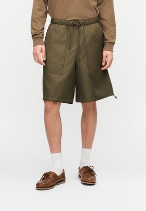 Homme portant un short kaki à cordon de serrage, une chemise marron à manches longues, des chaussettes blanches et des chaussures bateau en cuir marron, debout devant un fond uni.