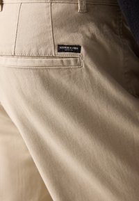 Beige cotton trousers with a smooth texture, featuring a rear pocket and a branded label in black and white.