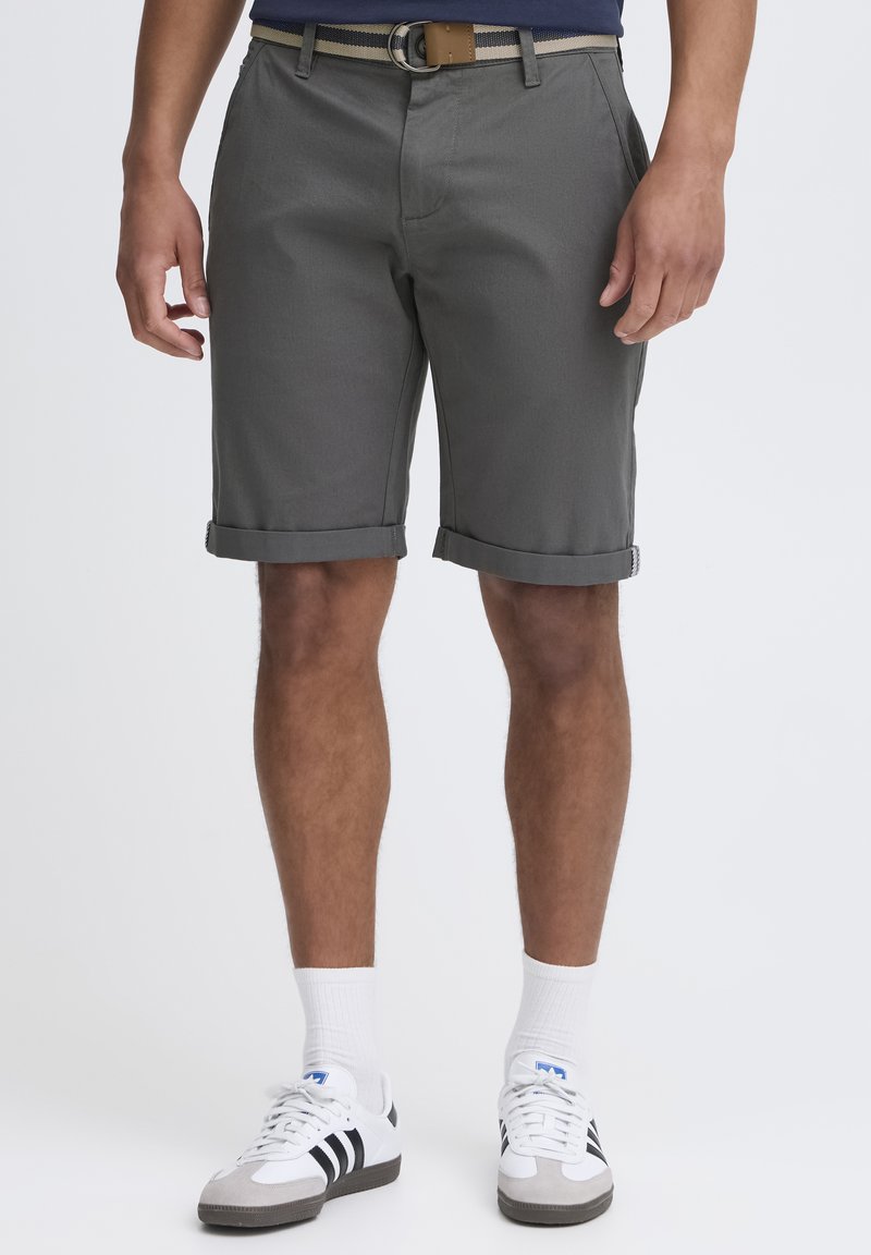 Pantalones cortos grises hasta la rodilla con puños, confeccionados en una tela ligera. Estilizados con un cinturón a rayas marrón y beige y zapatillas blancas.