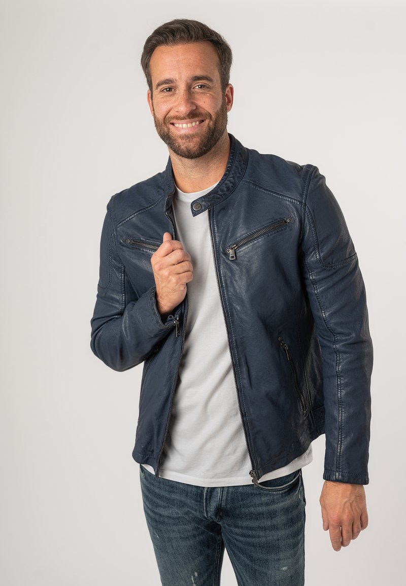 Giacca in pelle blu navy con colletto alto, dotata di tasche con zip e una vestibilità aderente. Abbinata a una camicia bianca e jeans.