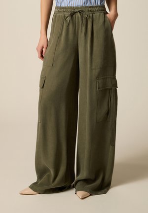 Olivengrønne cargo-bukser med vide ben, snøre i taljen, lappelommer og klaplommer, båret med beige spidse sko.