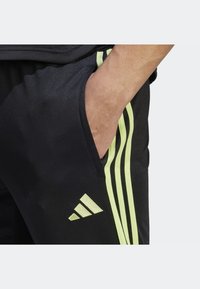 Černé sportovní kalhoty z hladké látky, s třemi svislými limetkově zelenými pruhy a kapsou na zip s logem Adidas.