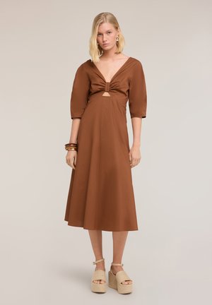 Mujer rubia que lleva un vestido midi marrón con mangas abullonadas y un nudo delantero, combinado con sandalias de plataforma beige y pulseras.