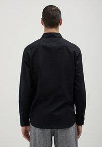 Chemise noire à manches longues avec col, présentant une texture lisse et des poignets boutonnés. Portée avec un pantalon léger gris, à motifs.