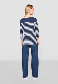 Tamsiai mėlynas ir baltas dryžuotas ilgomis rankovėmis marškinėliai su plačia apykakle, derinami su mėlynomis plačiomis kelnėmis ir baltais sportbačiais. Vientisas fonas.