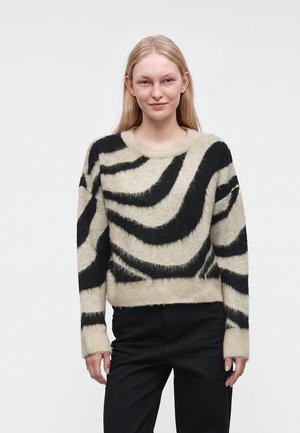Ung kvinde med glat blondt hår iført en beige og sort stribet blød sweater og sorte bukser, stående mod en ensfarvet baggrund.