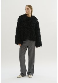 Zwarte, gestructureerde furjas met een oversized silhouet, gecombineerd met grijze broek met streepjes en crèmekleurige instapschoenen. Eenvoudige achtergrond.