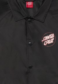 Svart vattentät jacka med knäppning framtill, meshlining och röd "Santa Cruz"-logotyp i en cirkulär design på vänster bröst.