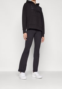 Personne portant un sweat à capuche noir, un pantalon ajusté noir et des baskets blanches, debout devant un fond clair uni.