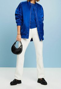 Blaue Satin-Bomberjacke über einem blauen Oberteil, weiße ausgestellte Jeans und schwarze Plateau-Loafers; trägt eine runde schwarze Handtasche.