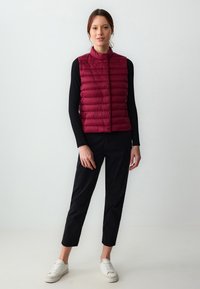 Gilet leggero color bordeaux imbottito con cuciture orizzontali, colletto alto e tasche con zip, abbinato a pantaloni neri e sneakers bianche.