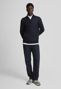 Pullover blu navy con zip e una texture sottile, abbinato a pantaloni coordinati e sneakers grigie. Il colletto si poggia su una camicia bianca.