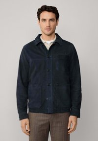 Marineblauwe suède jas met een kraag, voorknopsluiting, twee grote voortassen en een borstzak, met een soepele textuur.