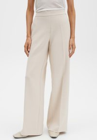 Beige broek met wijde pijpen en een gladde textuur, een hoge tailleband en verticale naaddetails; gecombineerd met een strak wit topje en lichtgekleurde schoenen.