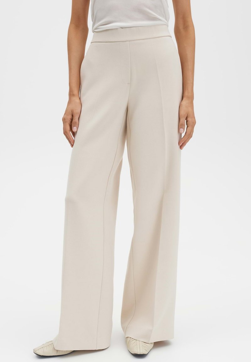 Beige broek met wijde pijpen en een gladde textuur, een hoge tailleband en verticale naaddetails; gecombineerd met een strak wit topje en lichtgekleurde schoenen.