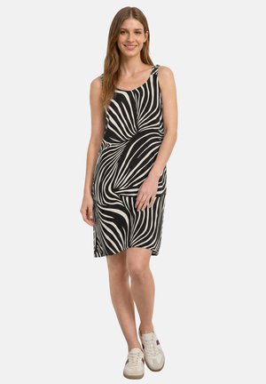 Femme aux longs cheveux portant une robe noire et blanche à motif tourbillonnant, sans manches, longueur genoux, et des baskets blanches, souriante avec une main sur la cuisse.