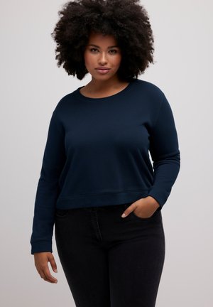 Sudadera recortada de color azul marino en material suave, con escote redondo y mangas largas, combinada con jeans negros. Presenta un ajuste relajado.
