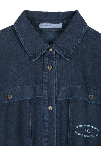 Chemise en denim foncé avec col et boutons, comportant un petit texte blanc brodé sur la poitrine qui dit "LET THE MYSTERY FIND YOU B.C."