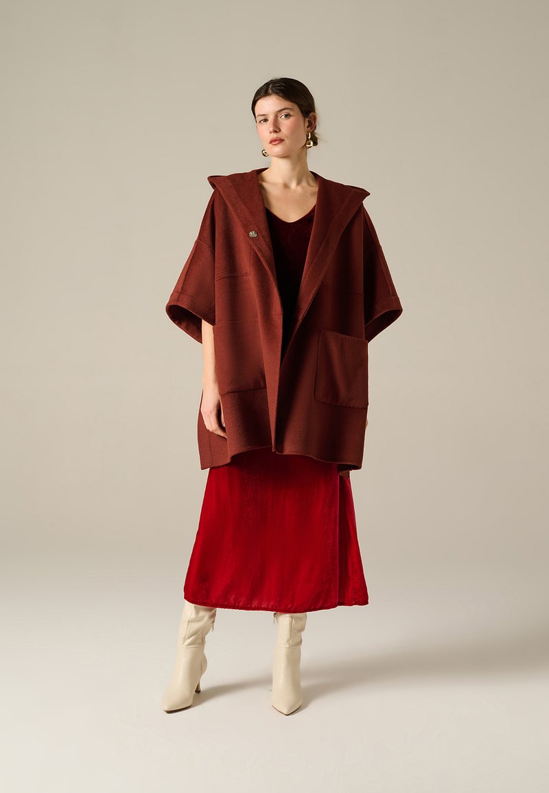 Cappotto chunky di colore ruggine con cappuccio, dotato di due grandi tasche e un bottone, indossato sopra un abito di velluto rosso a lunghezza ginocchio e stivali beige.