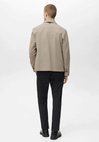 Beige jas met een klassieke kraag, zijzakken en een ontspannen pasvorm, gecombineerd met zwarte broek en donkere schoenen, gezien vanuit de achterkant.