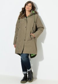 Cappotto lungo isolato verde oliva con cappuccio, corde di regolazione, tasche laterali e una fodera interna a contrasto. Indossato sopra un paio di jeans e stivali.