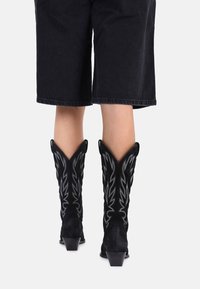 Sorte semskede cowboystøvler med spidse tæer, dekorativ hvid syning og en kort blokhæle, båret med mørke, knælange shorts.