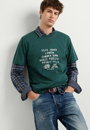 Giovane uomo con una t-shirt verde Pepe Jeans London del festival musicale sopra una camicia a quadri blu, abbinata a jeans blu slavati e una cintura marrone.