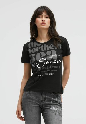 Young woman wearing a black graphic t-shirt and gray jeans with text prints, standing against a plain light background.