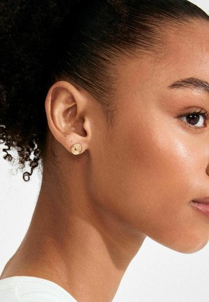 Zelta apaļa kniedes auskaru ar spirālveida dizainu, attēlots tuvinātā skata uz gludu ādu ar tumšiem, cirtainiem matiem.