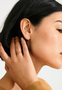 Anillo de oro con un pequeño acento verde, llevado en un dedo, junto a piel suave y cabello oscuro. Vista de perfil que resalta el diseño del anillo.