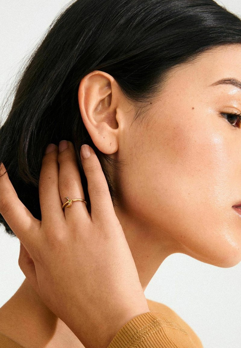 Anillo de oro con un pequeño acento verde, llevado en un dedo, junto a piel suave y cabello oscuro. Vista de perfil que resalta el diseño del anillo.