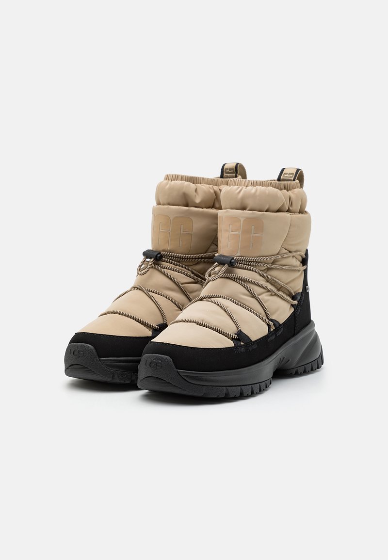 Beige und schwarze isolierte Winterstiefel mit dicken Sohlen und elastischen Bungee-Schnürsenkeln, Logo im gepolsterten Knöchelbereich und vorne an der Zehenbox geprägt.