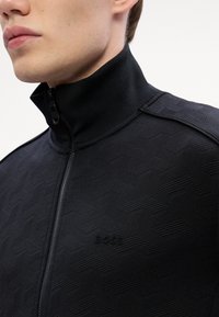 Giacca nera con zip e colletto alto, caratterizzata da un motivo geometrico testurizzato. Logo "BOSS" in rilievo sul lato sinistro.