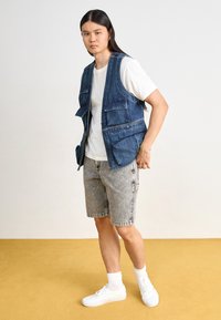 Personne portant un t-shirt blanc, un gilet en jean bleu avec plusieurs poches, un short en jean gris, des chaussettes blanches et des baskets blanches debout sur un sol jaune.