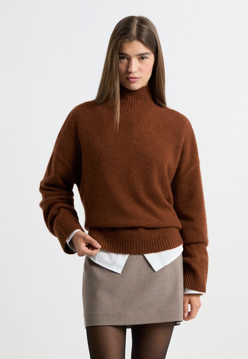 Brązowy sweter z dzianiny z wysokim kołnierzem, prążkowanym wykończeniem i oversize'owymi rękawami, w połączeniu z jasną koszulą i szarą spódnicą w wzory.