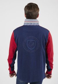 Chemise à manches longues bleu marine et rouge avec col à motifs. Présente un logo brodé au dos. Texture lisse, coupe décontractée et style casual.