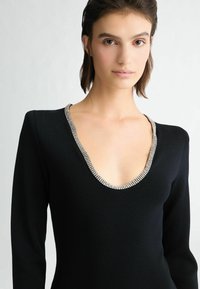 Schwarzes Kleid mit tiefem V-Ausschnitt, verziert mit silberfarbenen Strasssteinen, langen, figurbetonten Ärmeln und einer glatten Textur.