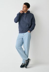 Tamsiai mėlynas džemperis su gobtuvu, kišene priekyje, raišteliais ir ribotais rankogaliais. Derinamas su šviesiai mėlynomis, tiesiomis džinsų kelnėmis ir juodais New Balance sportbačiais.