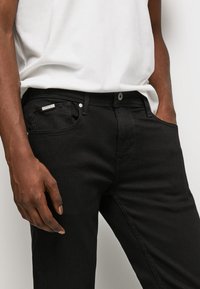 Jeans de mezclilla negros de corte ajustado, con un bolsillo frontal, herrajes plateados y costuras en contraste. Llevados con una camiseta blanca sencilla.