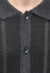 Tamnosiva pletena jakna s teksturiranim završetkom, s ovratnikom i istaknutim kopčanjem na gumbe. Suptilne pruge u različitim nijansama.