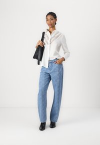Camicia bianca con dettagli in pizzo lungo il colletto, abbinata a jeans leggere blu con fantasia e stivali neri a caviglia, mentre tiene una borsa nera.